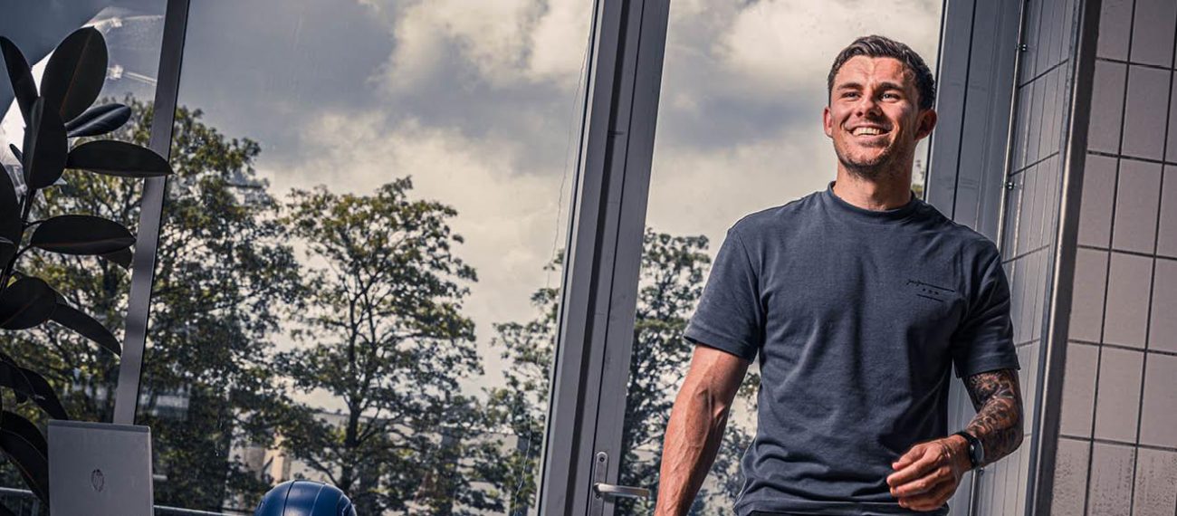 Lachende persoon in casual kleding lopend door een industriële binnenruimte met grote ramen en planten op de achtergrond