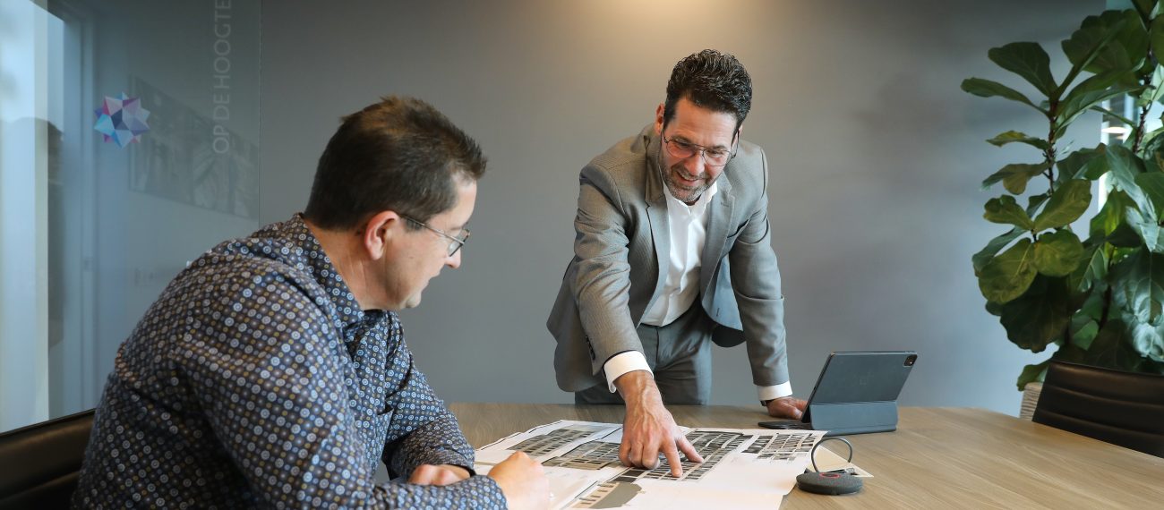 Twee mannen aan een vergadertafel bekijken bouwtekeningen, terwijl één van hen wijst en toelichting geeft.