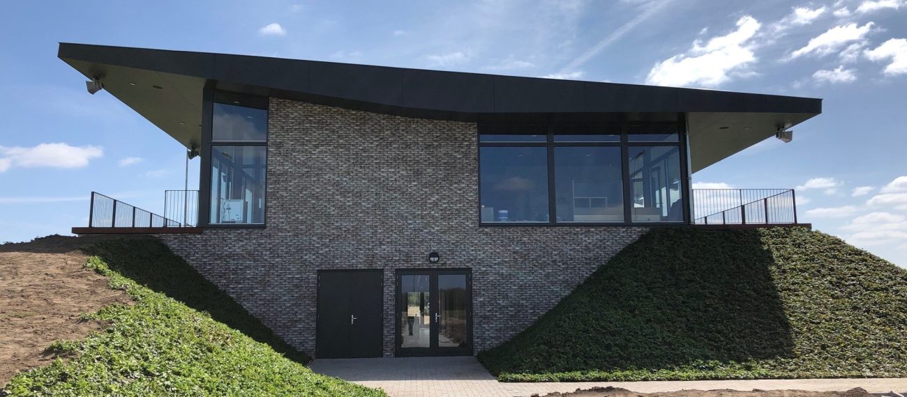 Voorzijde van het nieuwe clubgebouw DVO’20 met uitkragend dak, grote glaspuien en met klimop begroeide taluds; een klinkerpad leidt naar de entree in de bakstenen gevel onder een blauwe lucht.
