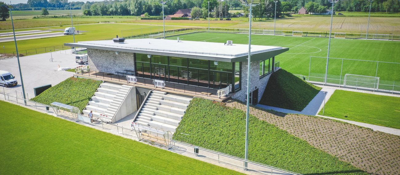 Luchtfoto van het moderne clubgebouw van DVO’20 in Kelpen-Oler, met glazen pui, tribunetrap en groene taluds, omgeven door meerdere voetbalvelden.