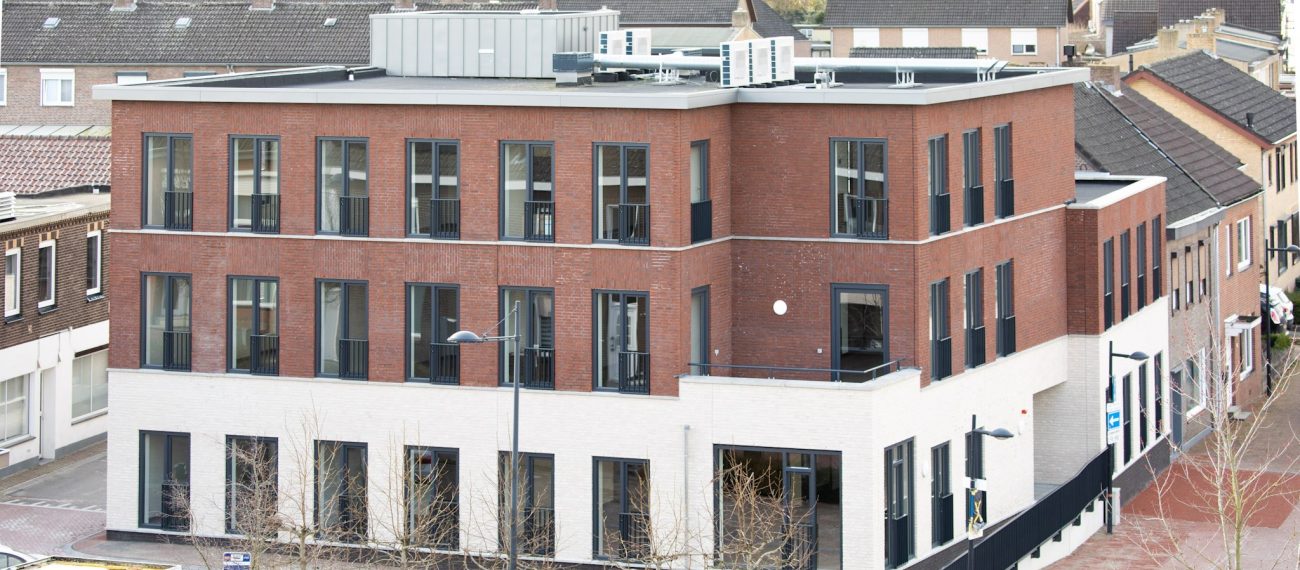 Nieuwbouw zorgcomplex Cicero Bleekstaete in Schinveld: modern hoekgebouw van drie verdiepingen met rood bakstenen gevel en witte plint, veel hoge ramen en een plat dak met installaties; gelegen aan een straat met jonge, bladloze bomen en een bushalte, omringd door woonhuizen.