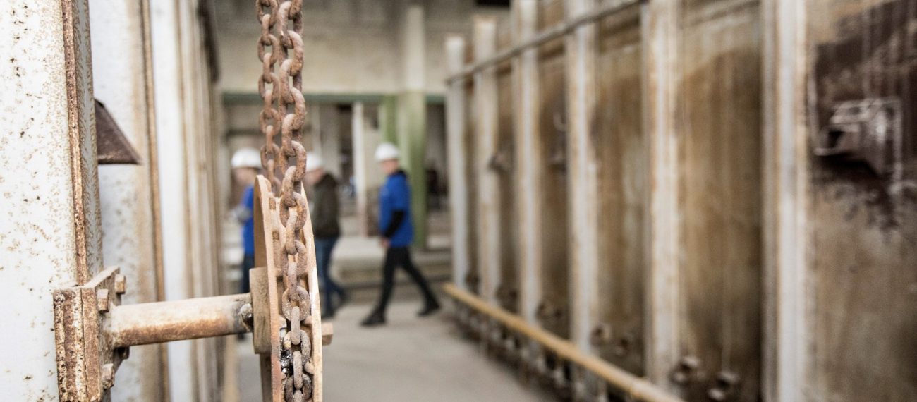 Close-up van een roestige ketting en tandwiel in een oude industriële ruimte, met twee personen met bouwhelmen wazig op de achtergrond.
