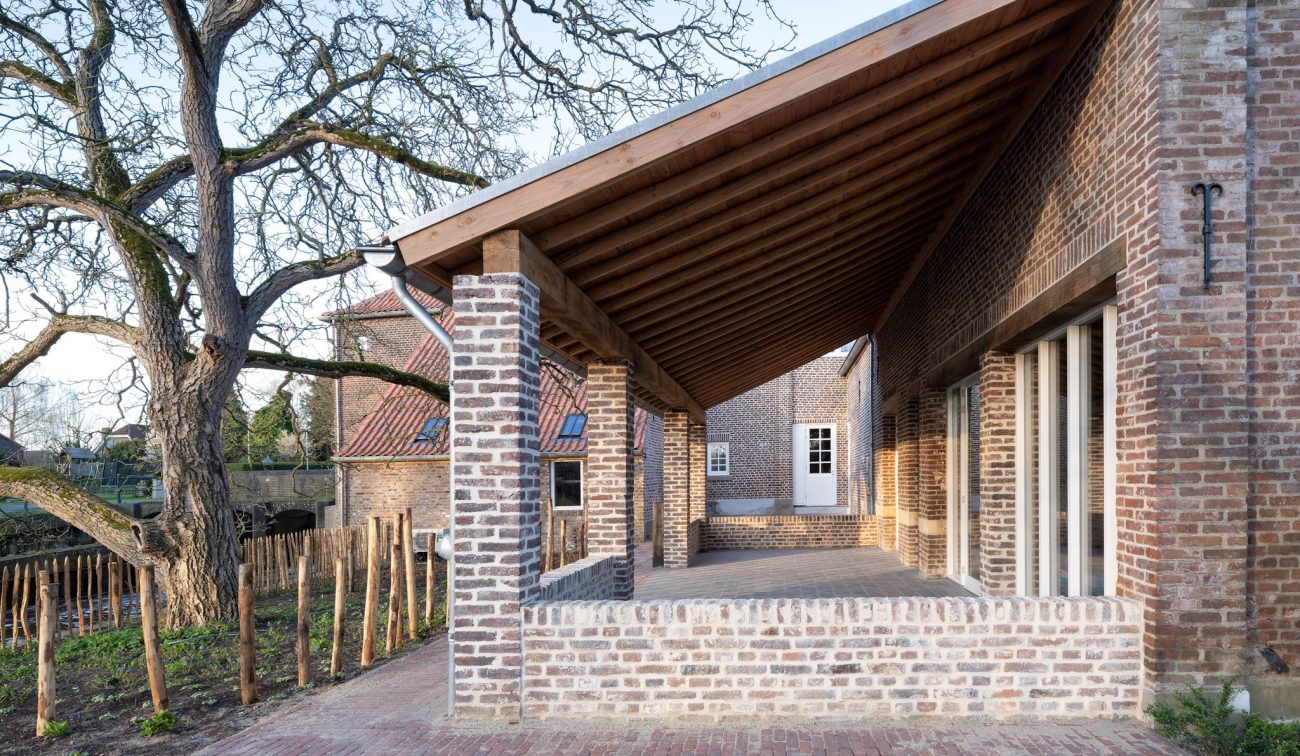 Overdekte bakstenen veranda met houten spanten en pilaren, uitkijkend op de binnenplaats van de gerestaureerde Grathemermolen en een grote boom.
