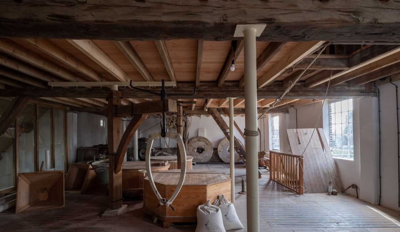 Interieur Grathemermolen met houten balken en vloeren, historische maalinstallatie met ronde maalstenen, hijsring en graanzakken, verlicht door hoge roedeverdeelde ramen.