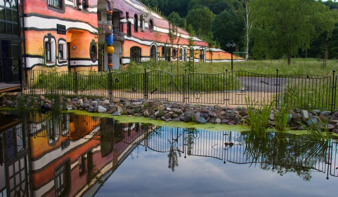 Ronald McDonald Kindervallei in Valkenburg met golvende, kleurrijke gevel die weerspiegelt in een vijver, omgeven door groen en hekwerk.