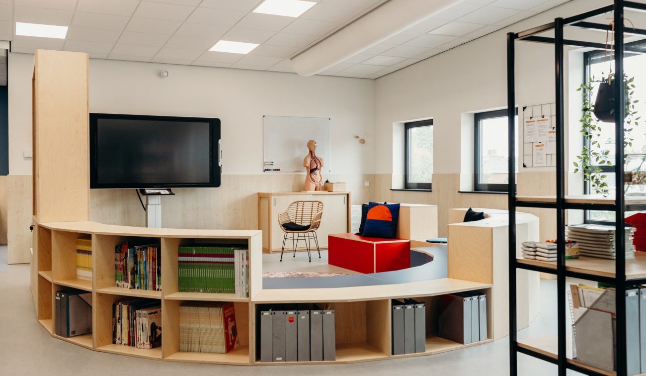 Moderne klasruimte met gebogen houten boekenkast als zittribune, interactieve scherm en whiteboard, anatomiemodel op kast, ramen met daglicht, en modulaire zitblokken met kussens; rechts een open stellingkast met schriften.