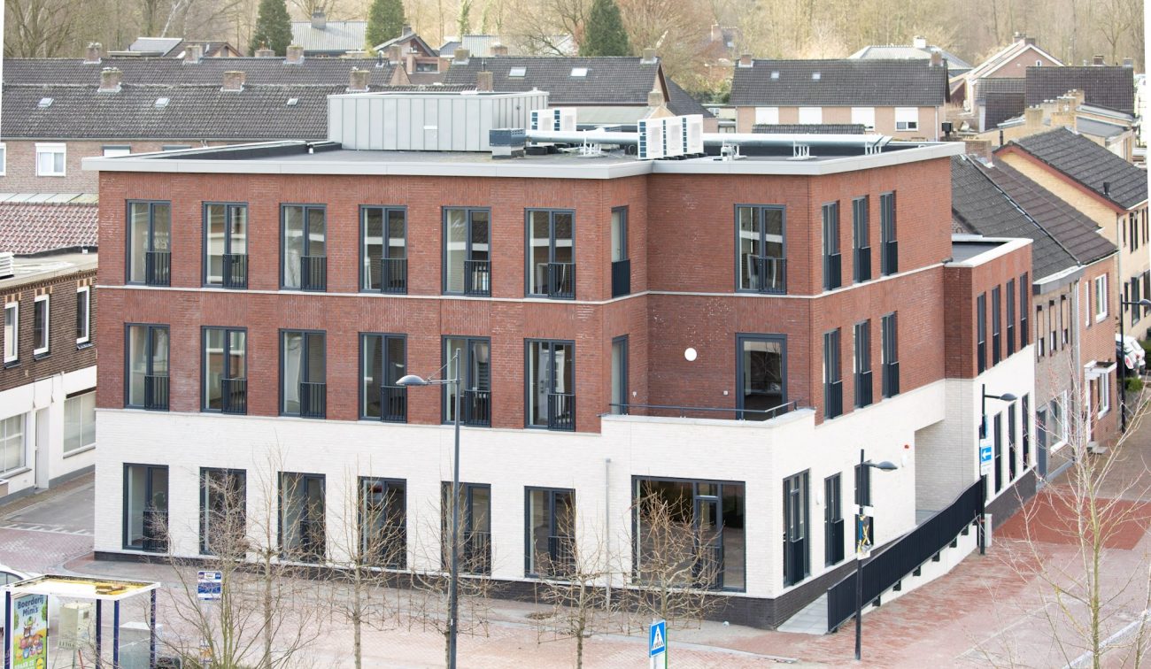 Nieuwbouw zorgcomplex Cicero Bleekstaete in Schinveld: modern hoekgebouw van drie verdiepingen met rood bakstenen gevel en witte plint, veel hoge ramen en een plat dak met installaties; gelegen aan een straat met jonge, bladloze bomen en een bushalte, omringd door woonhuizen.