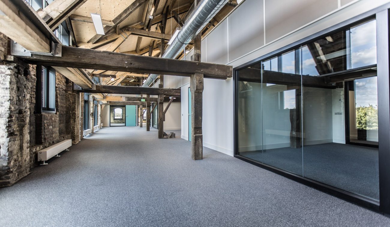 Industriële gang met zichtbare houten balken en bakstenen wanden, glazen kantoorruimtes rechts, zichtbare luchtkanalen onder het schuine dak en grijze tapijtvloer.