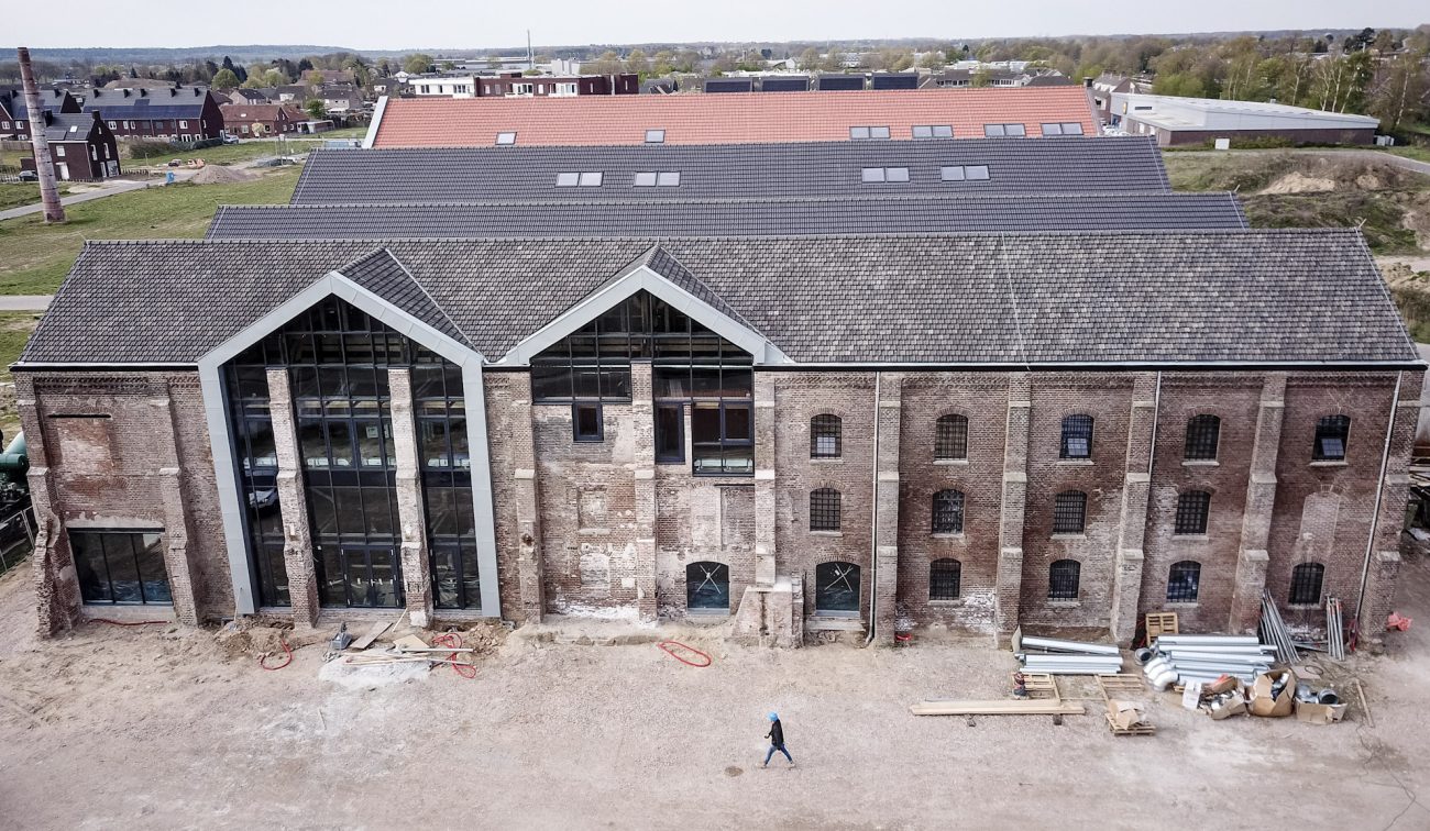 Bakstenen gevel van de Greswarenfabriek in Reuver in renovatie, met grote glazen topgevels; op de voorgrond bouwterrein met materialen en een passerende bouwmedewerker.”