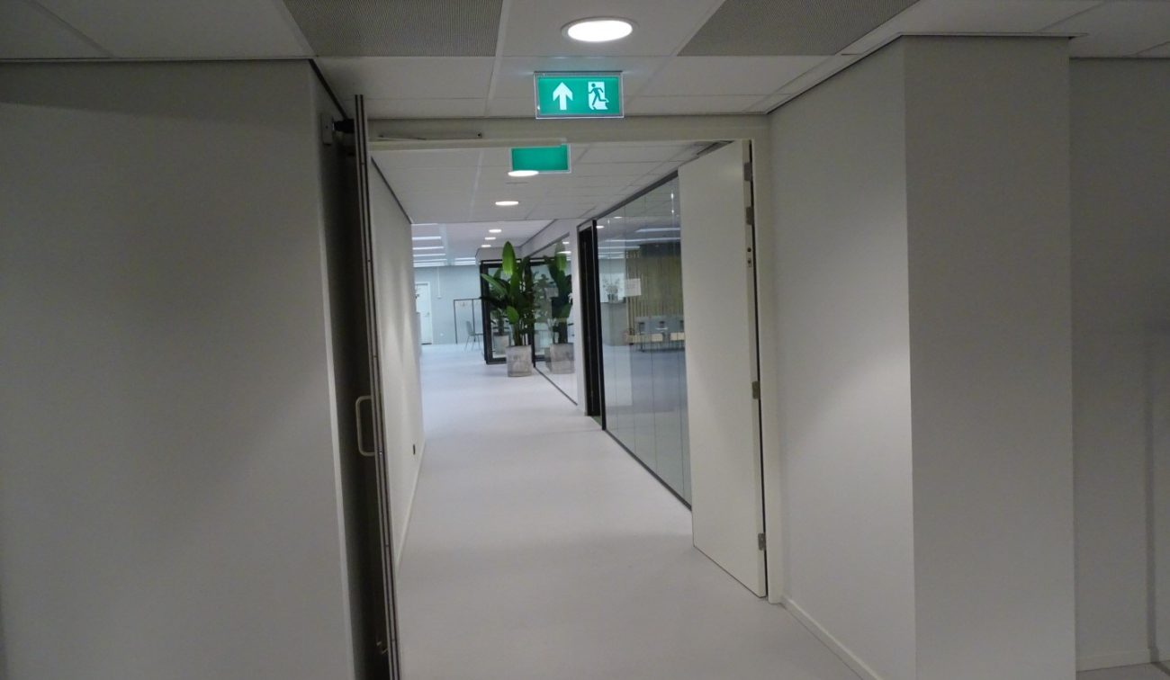 Gang Meeting Plaza: witte wanden, glazen kantoorwand, open deur en groen nooduitgangsbord bovenin