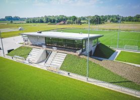 Luchtfoto van het moderne clubgebouw van DVO’20 in Kelpen-Oler, met glazen pui, tribunetrap en groene taluds, omgeven door meerdere voetbalvelden.