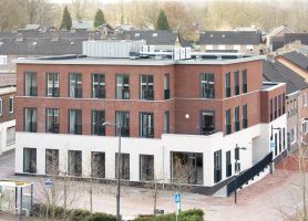 Nieuwbouw zorgcomplex Cicero Bleekstaete in Schinveld: modern hoekgebouw van drie verdiepingen met rood bakstenen gevel en witte plint, veel hoge ramen en een plat dak met installaties; gelegen aan een straat met jonge, bladloze bomen en een bushalte, omringd door woonhuizen.