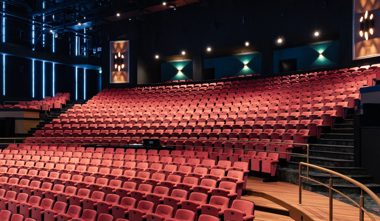 Grote theaterzaal van De Oranjerie met oplopende rijen rode stoelen, houten looppaden langs de zijkant en donkere wanden met verticale LED-lijnen.