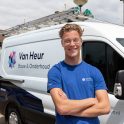 Een jonge medewerker staat glimlachend met gekruiste armen voor een bedrijfsbus met het logo ‘Van Heur Bouw & Onderhoud’