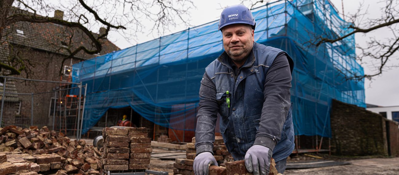 Metselaar met bouwhelm sorteert oude bakstenen op een bouwplaats met steigers en blauwe afscherming op de achtergrond.