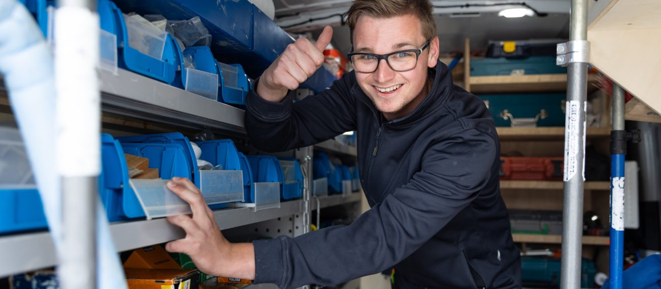 Lachende medewerker met bril die in een busje tussen opbergbakken staat en duim omhoog steekt.