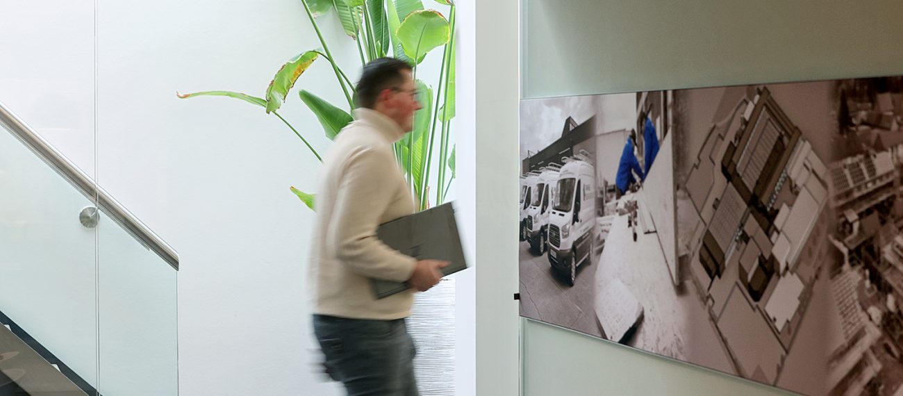 Medewerker loopt door modern kantoor langs wandfoto met bestelwagens en bouwproject, met grote planten