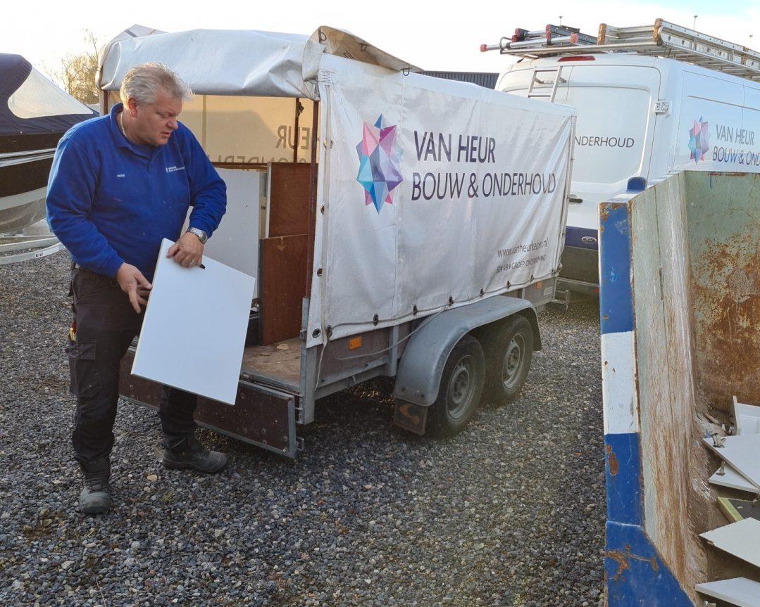 Medewerker van Van Heur Bouw & Onderhoud draagt plaatmateriaal naast aanhanger en bouwafvalcontainer