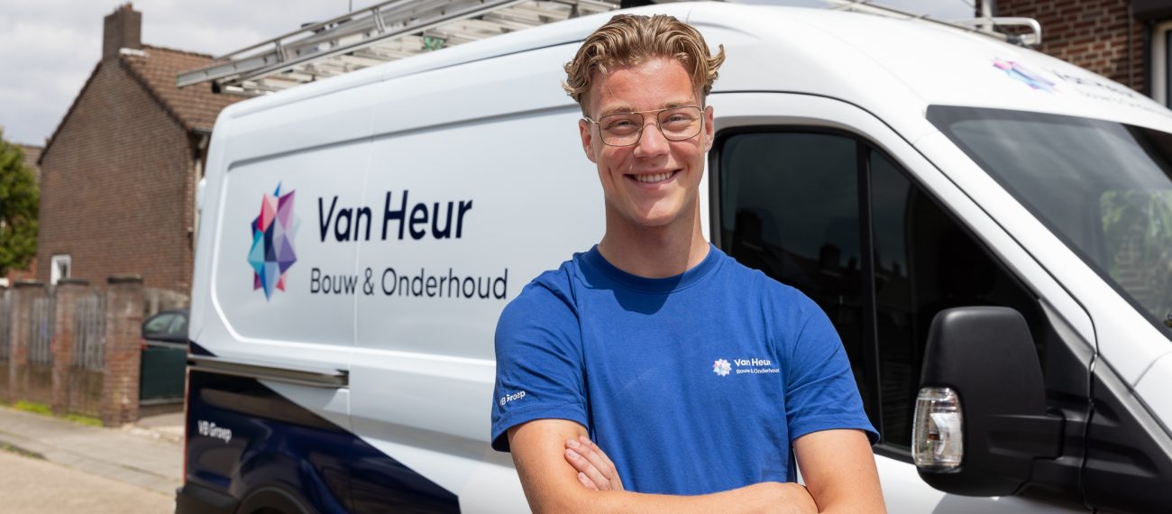 Een jonge medewerker staat glimlachend met gekruiste armen voor een bedrijfsbus met het logo ‘Van Heur Bouw & Onderhoud’