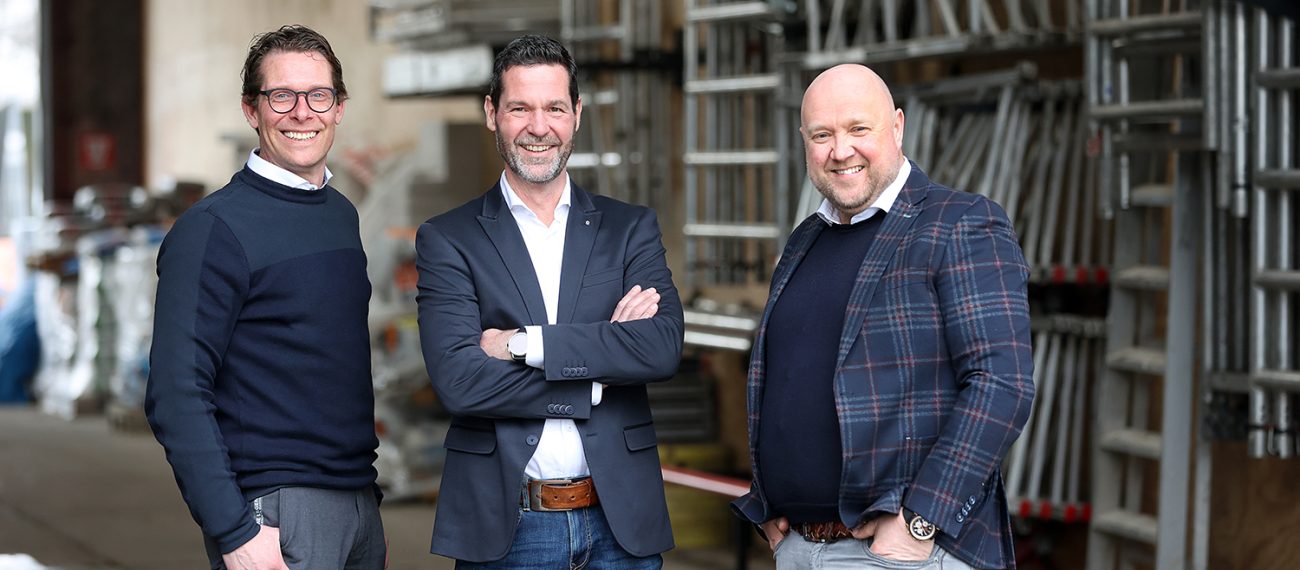 Drie mannen in nette kleding staan lachend voor een opslagruimte met stapels steigermateriaal.