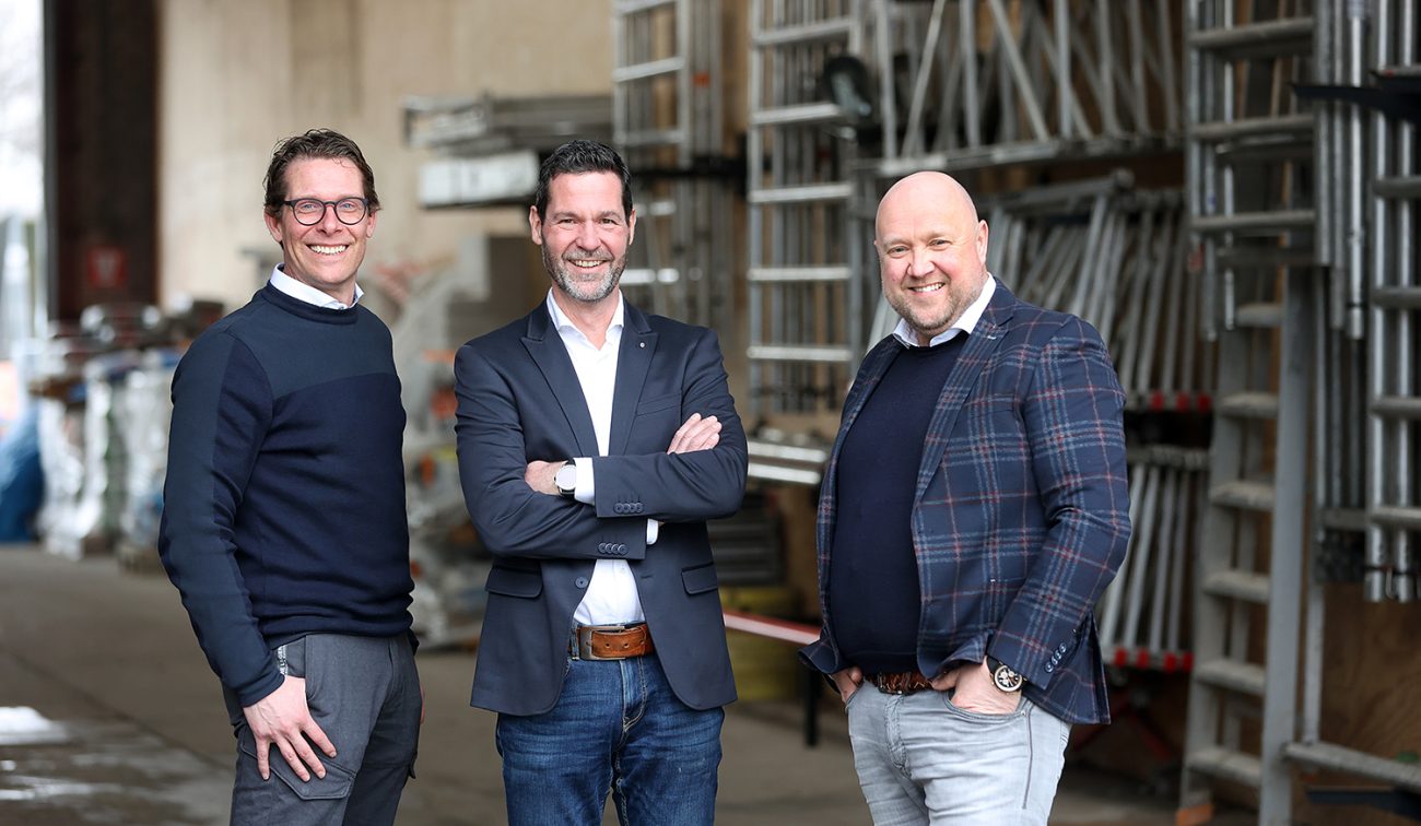 Drie mannen in nette kleding staan lachend voor een opslagruimte met stapels steigermateriaal.