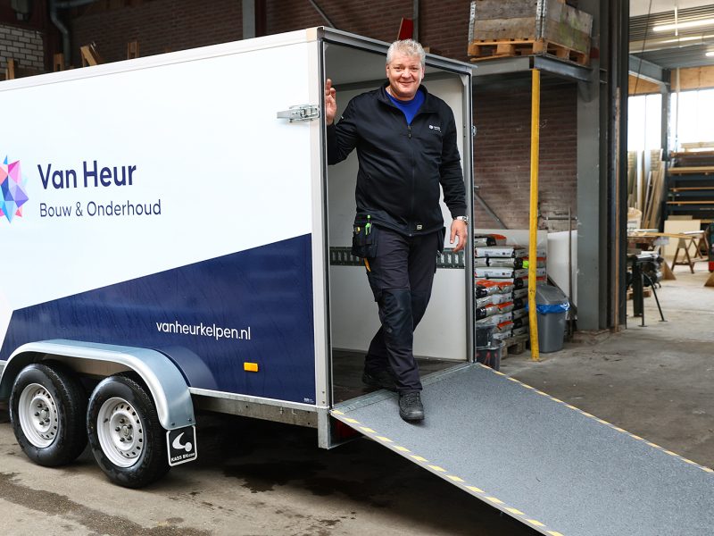 Medewerker stapt uit bedrijfsaanhanger met oprijplaat in werkplaatsomgeving, met logo van Van Heur Bouw & Onderhoud op de zijkant.”