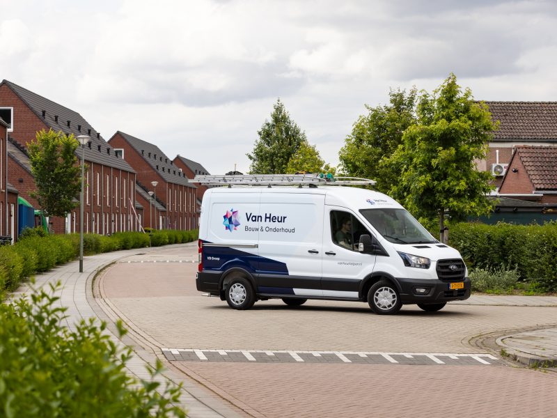 Bedrijfsbus van Van Heur Bouw & Onderhoud rijdt door een woonwijk met nieuwbouwwoningen.