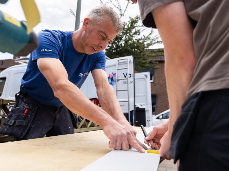 Bouwmedewerker meet en tekent af op houtplaat tijdens buitenwerk, met bedrijfsbus op de achtergrond.