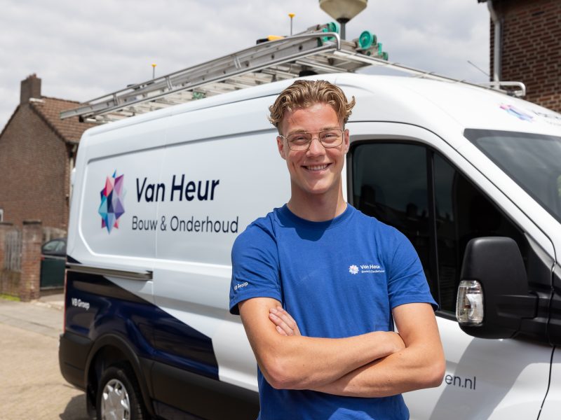Een jonge medewerker staat glimlachend met gekruiste armen voor een bedrijfsbus met het logo ‘Van Heur Bouw & Onderhoud’