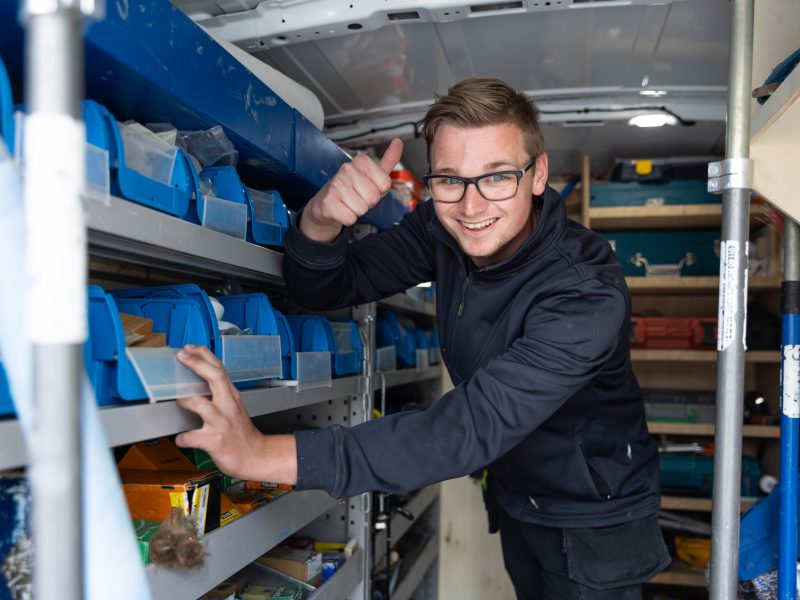 Lachende medewerker met bril die in een busje tussen opbergbakken staat en duim omhoog steekt.