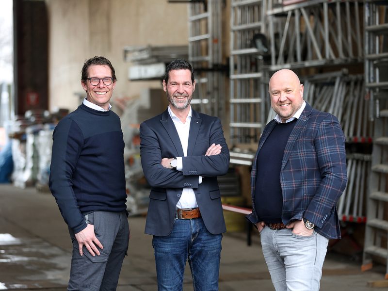 Drie mannen in nette kleding staan lachend voor een opslagruimte met stapels steigermateriaal.