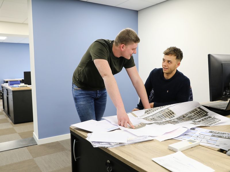 Twee mannen bespreken bouwtekeningen aan een bureau in een kantooromgeving.
