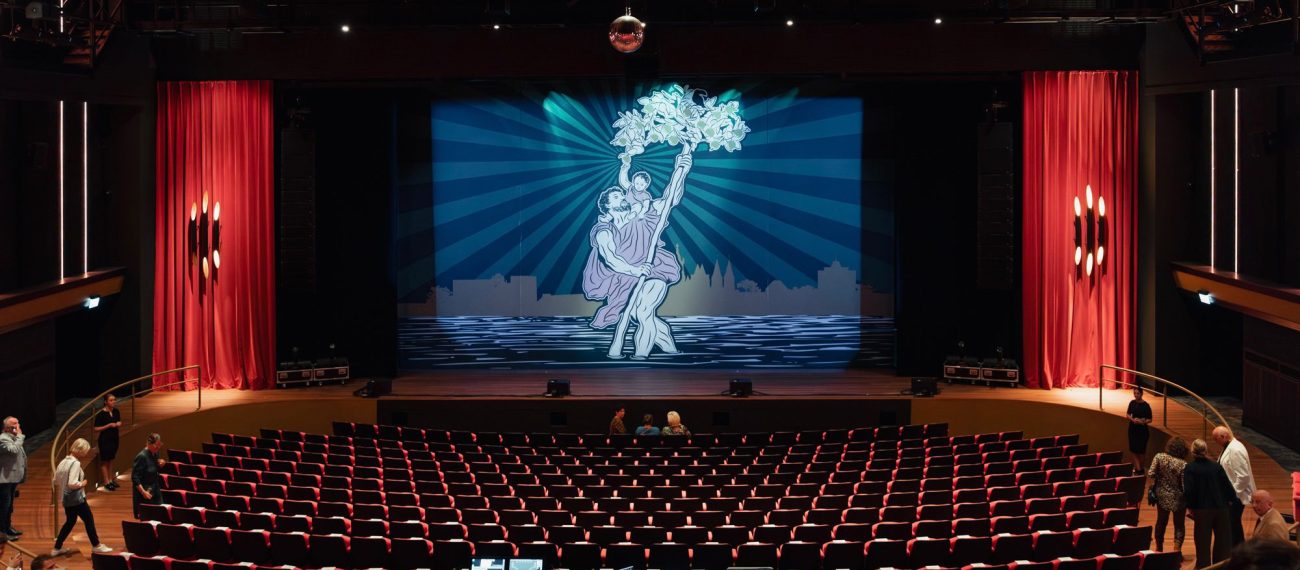 Theaterzaal met rode stoelen en zicht op het podium met projectie van een dansend paar; techniektafel centraal en enkele bezoekers die hun plaatsen innemen.