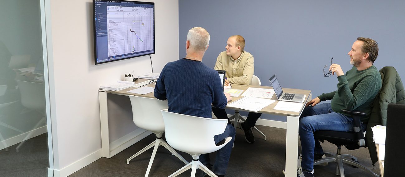 Drie medewerkers zitten aan een vergadertafel en bespreken een planning die op een groot scherm aan de muur wordt weergegeven.