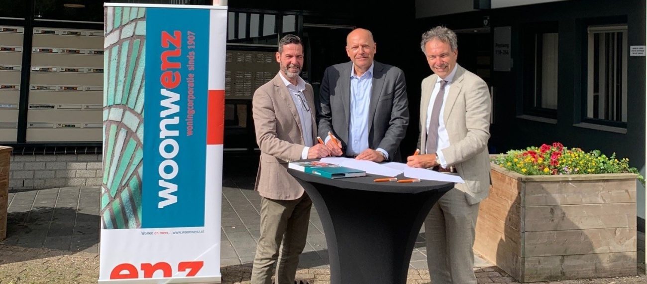 Drie mannen staan bij een statafel voor een gebouw met een banner en een plattegrond.