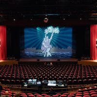 Theaterzaal De Oranjerie met rijen rode stoelen, een mengtafel achterin en een podium met rode zijgordijnen en projectie van een dansend paar met bloemen; bezoekers nemen plaats.