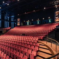 Grote theaterzaal van De Oranjerie met oplopende rijen rode stoelen, houten looppaden langs de zijkant en donkere wanden met verticale LED-lijnen.