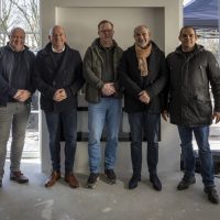 Vijf mannen poseren samen in een lichte ruimte met ramen op de achtergrond.