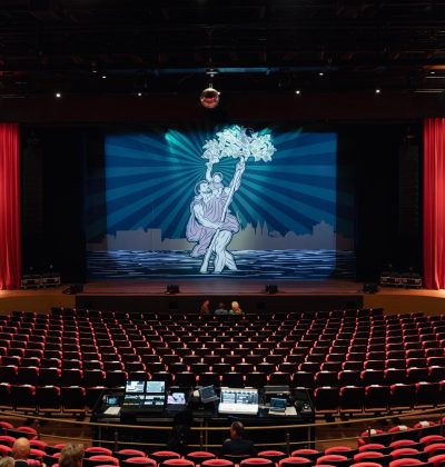 Theaterzaal met rode stoelen en zicht op het podium met projectie van een dansend paar; techniektafel centraal en enkele bezoekers die hun plaatsen innemen.