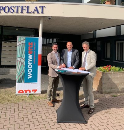 Drie mannen staan bij een statafel voor een gebouw met een banner en een plattegrond.