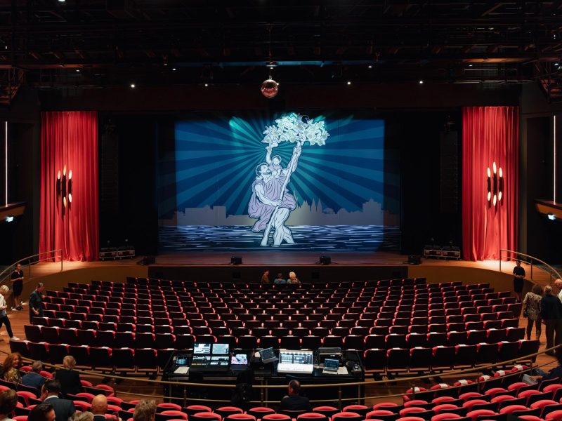 Theaterzaal De Oranjerie met rijen rode stoelen, een mengtafel achterin en een podium met rode zijgordijnen en projectie van een dansend paar met bloemen; bezoekers nemen plaats.