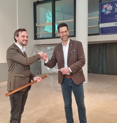 Twee mannen verrichten openingshandeling met schaar en rood lint in lichte zaal