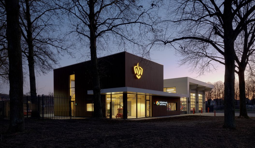 Moderne brandweerpost Sittard-Oost in de schemering met verlichte gevel en glazen entree; rechts de uitrijhal voor voertuigen, omlijst door silhouetten van bomen.