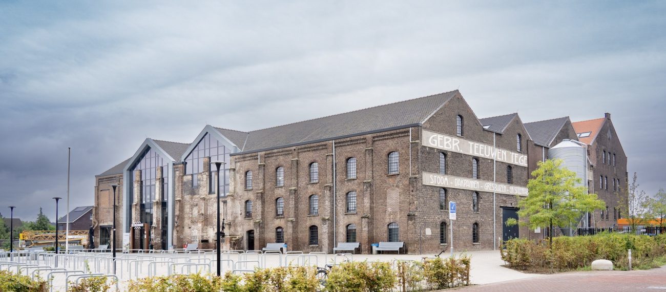 Gerestaureerde Greswarenfabriek in Reuver met bakstenen gevels, grote glazen entree en historische gevelletters; voorliggend plein met fietsrekken, bankjes en jonge bomen.