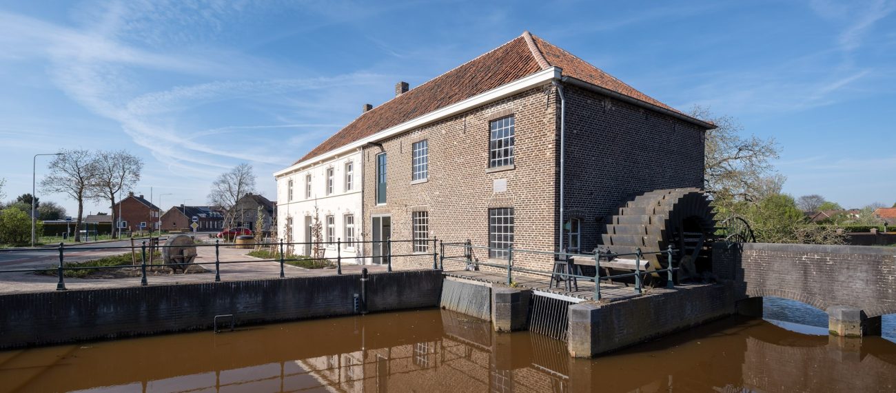 Grathemermolen in Grathem – bakstenen watermolen met rood pannendak en groot waterrad aan het water, met spiegeling in de molenvijver onder blauwe lucht.