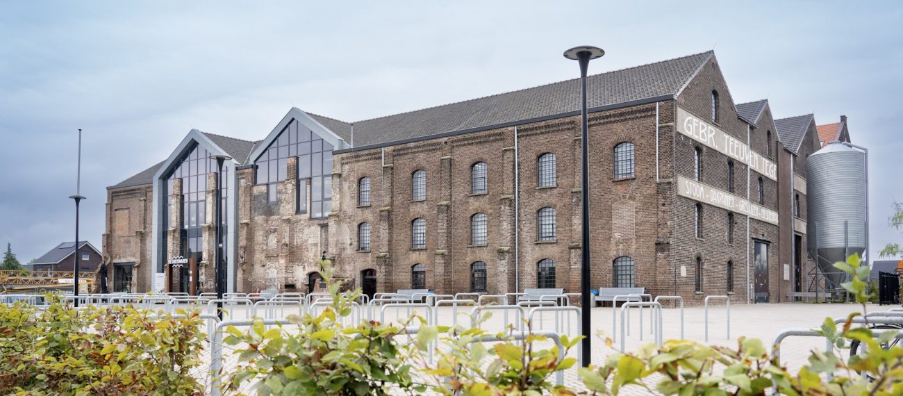 Getransformeerde Greswarenfabriek in Reuver: gerestaureerd bakstenen fabriekspand met grote glazen entree, oude gevelletters en silo aan het plein met fietsenrekken en lantaarnpalen.