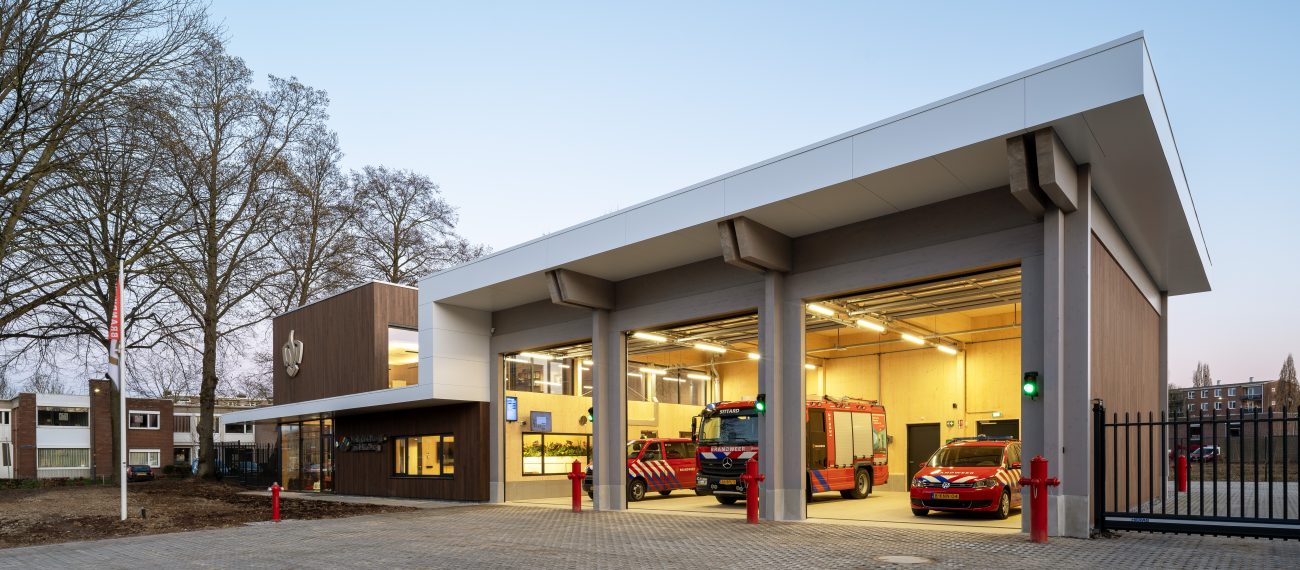 Nieuwe brandweerpost Sittard-Oost bij schemering met drie open uitrijpoorten; binnen staan een brandweerwagen en dienstauto klaar, naast de glazen entree in het moderne hout-wit gebouw.