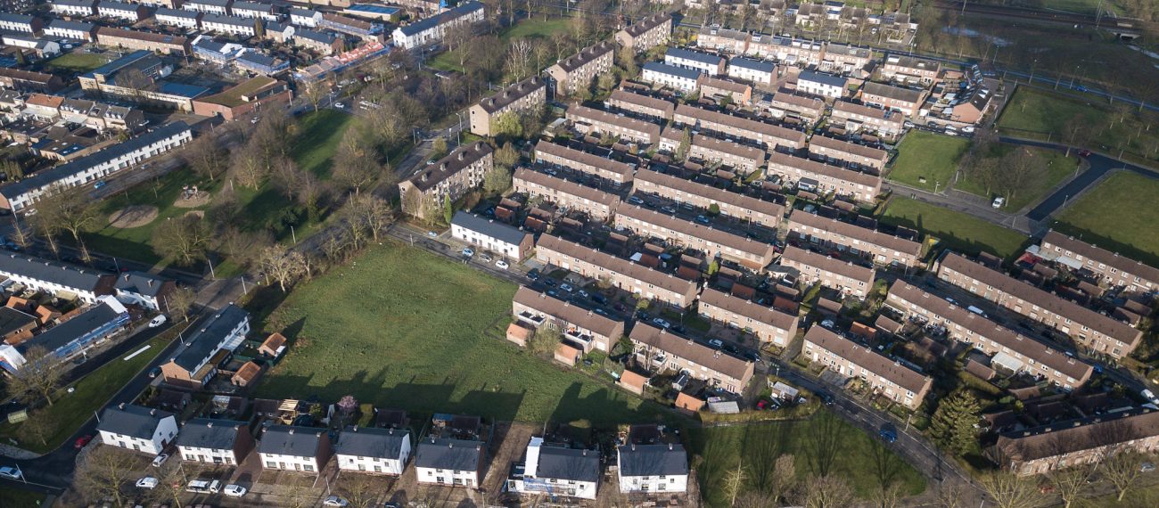 Luchtfoto van een woonwijk met lange rijen rijtjeshuizen en groene veldjes tussen de straten; renovatieblokken met lichte gevels vallen op tussen oudere baksteenwoningen.