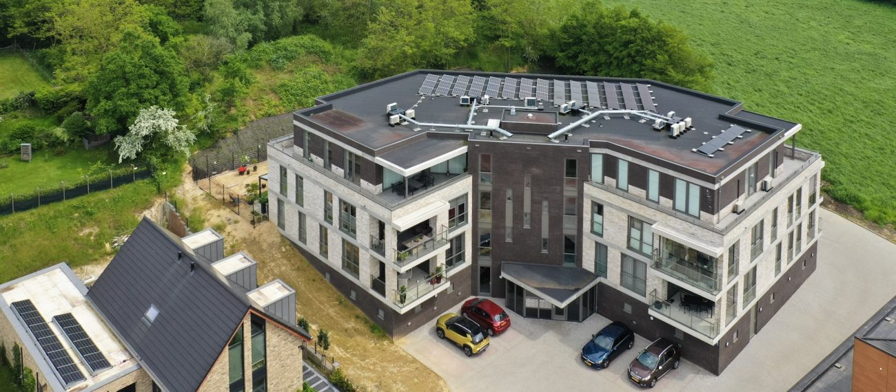 Schuin-van-bovenaanzicht van appartementengebouw Villa Rodendael in Vaesrade, met donker-plat dak vol zonnepanelen, gevels in donker baksteen en licht natuursteen, meerdere balkons en geparkeerde auto’s; omringd door groen en velden.