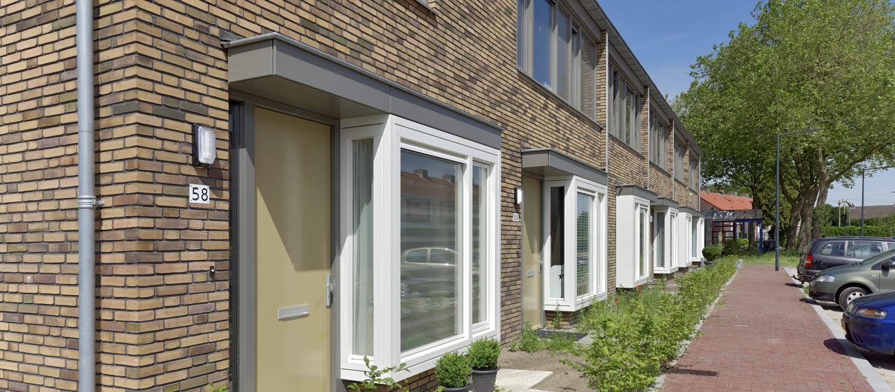 Rij van moderne eengezinswoningen met bakstenen gevels, witte erkers en beige voordeuren, gelegen aan een rustige straat met parkeerplaatsen en bomen.